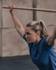 ELEIKO 木製テクニックバー ナチュラルウッド 150cm 女性が頭上で保持する動作を斜め前方から捉えた使用シーンでグリップ位置と長さ感が分かる