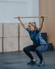 ELEIKO 木製テクニックバー ナチュラルウッド 150cm 女性がオーバーヘッドスクワットを行う全身構図でフォームとバーのサイズ感が分かるトレーニングシーン