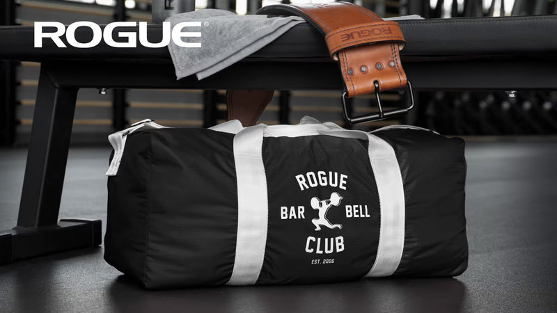 ROGUE オールドスクールジムバッグ ブラック、やや斜め前方からベンチ上に置いた状態を捉えた構図、ホワイトロゴプリントとグレーの持ち手ストラップ、円筒シルエットが分かる全体形状