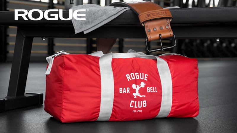 ROGUE オールドスクールジムバッグ レッド、やや斜め前方からベンチ上に置いた状態を捉えた構図、ロゴプリントとグレーの持ち手ストラップが確認できる全体デザイン