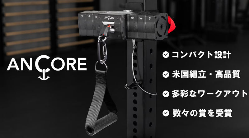 パワーラック支柱に装着したANCOREレジスタンスケーブル本体を斜め接写。黒マット仕上げのユニットと金属カラビナ付きハンドルが垂れ、右側にコンパクト設計・米国組立・多彩なワークアウト対応などの特長を白文字で示したジム内プロモーション構図。