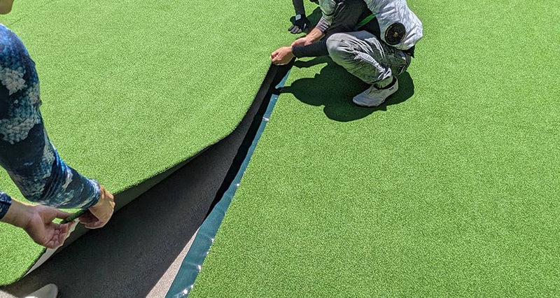 屋外施工現場で人工芝ロールを持ち上げ接合する作業を俯瞰で撮影、ジョイントテープを使った敷設工程の様子